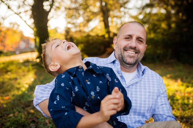 Little boy throwing his head back in laughter, sitting in his dad's lap.